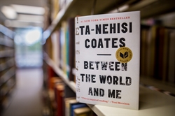 [ai] A book titled "Between the World and Me" by Ta-Nehisi Coates displayed on a shelf in a library. The cover features large black text on a white background with a circular award seal.